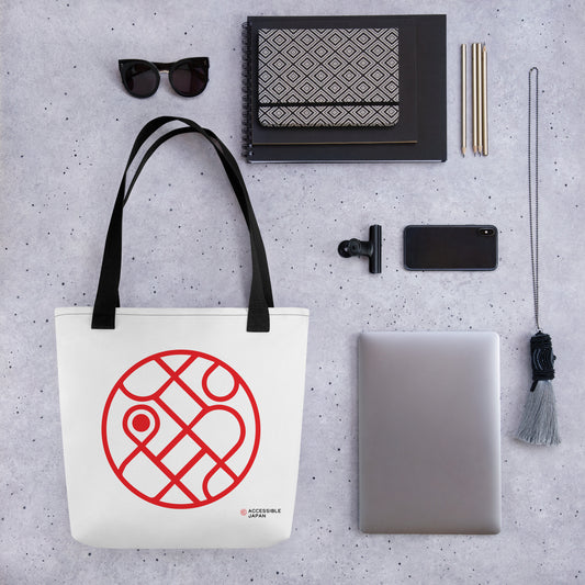 A white tote bag with black straps and a red circular design and the Accessible Japan logo, placed alongside sunglasses, notebooks, pencils, a phone, and a laptop.