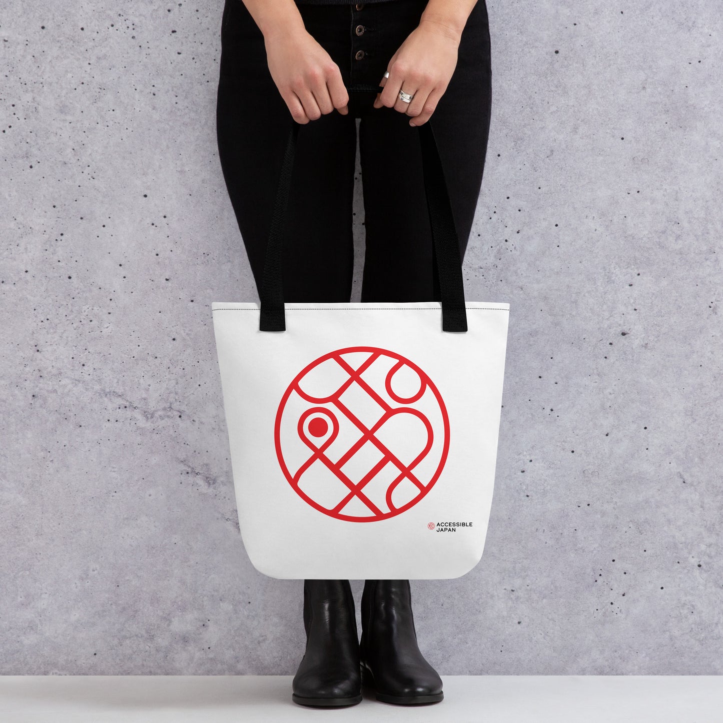 A person in black pants and boots holds a white tote bag with black straps, featuring a red circular design and the "Accessible Japan" logo.