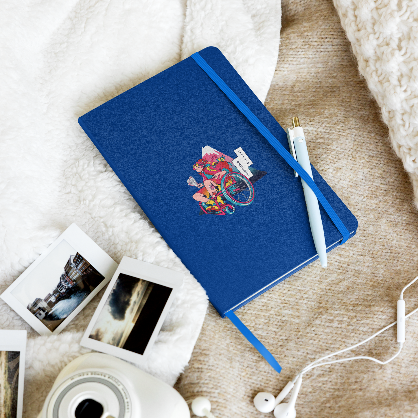 A blue notebook with a wheelchair traveler illustration lies on a cozy blanket, surrounded by instant photos, a pen, and earphones.