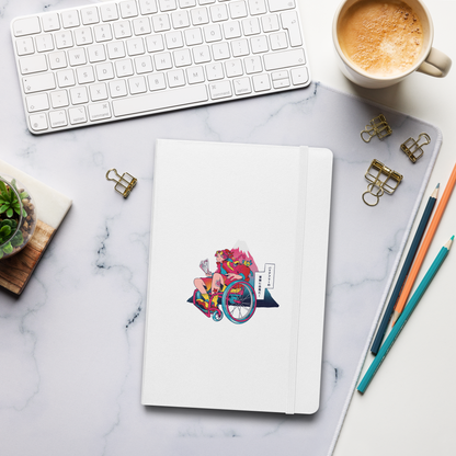 A white notebook with a colorful wheelchair traveler illustration lies on a marble desk, surrounded by a keyboard, coffee, and stationery.